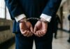 Miami Springs Resident and Coral Gables Lawyer Jackson Lainez Suspended by Florida Supreme Court Amid Lewd Conduct Charges Close-up of a man in a professional dark navy blue suit and white shirt cuffed in silver metal handcuffs behind his back.