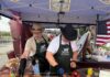 VGPD ‘Gettin’ Piggy Wit It’ Team Wins Best Decorated Booth at Blessed Trinity Holy Smokes Pig Roast Two men wearing cowboy hats and aprons prepare a whole roasted pig under a Virginia Gardens Police Department pop-up tent decorated with an American flag.