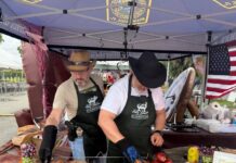VGPD ‘Gettin’ Piggy Wit It’ Team Wins Best Decorated Booth at Blessed Trinity Holy Smokes Pig Roast Two men wearing cowboy hats and aprons prepare a whole roasted pig under a Virginia Gardens Police Department pop-up tent decorated with an American flag.