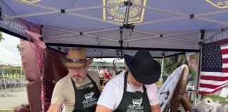 VGPD ‘Gettin’ Piggy Wit It’ Team Wins Best Decorated Booth at Blessed Trinity Holy Smokes Pig Roast Two men wearing cowboy hats and aprons prepare a whole roasted pig under a Virginia Gardens Police Department pop-up tent decorated with an American flag.